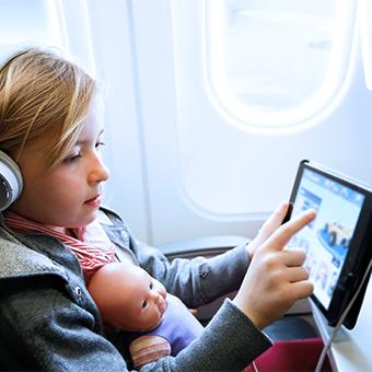Young girl wearing headphones with a baby doll in her lap playing a game on her tablet over inflight WiFi