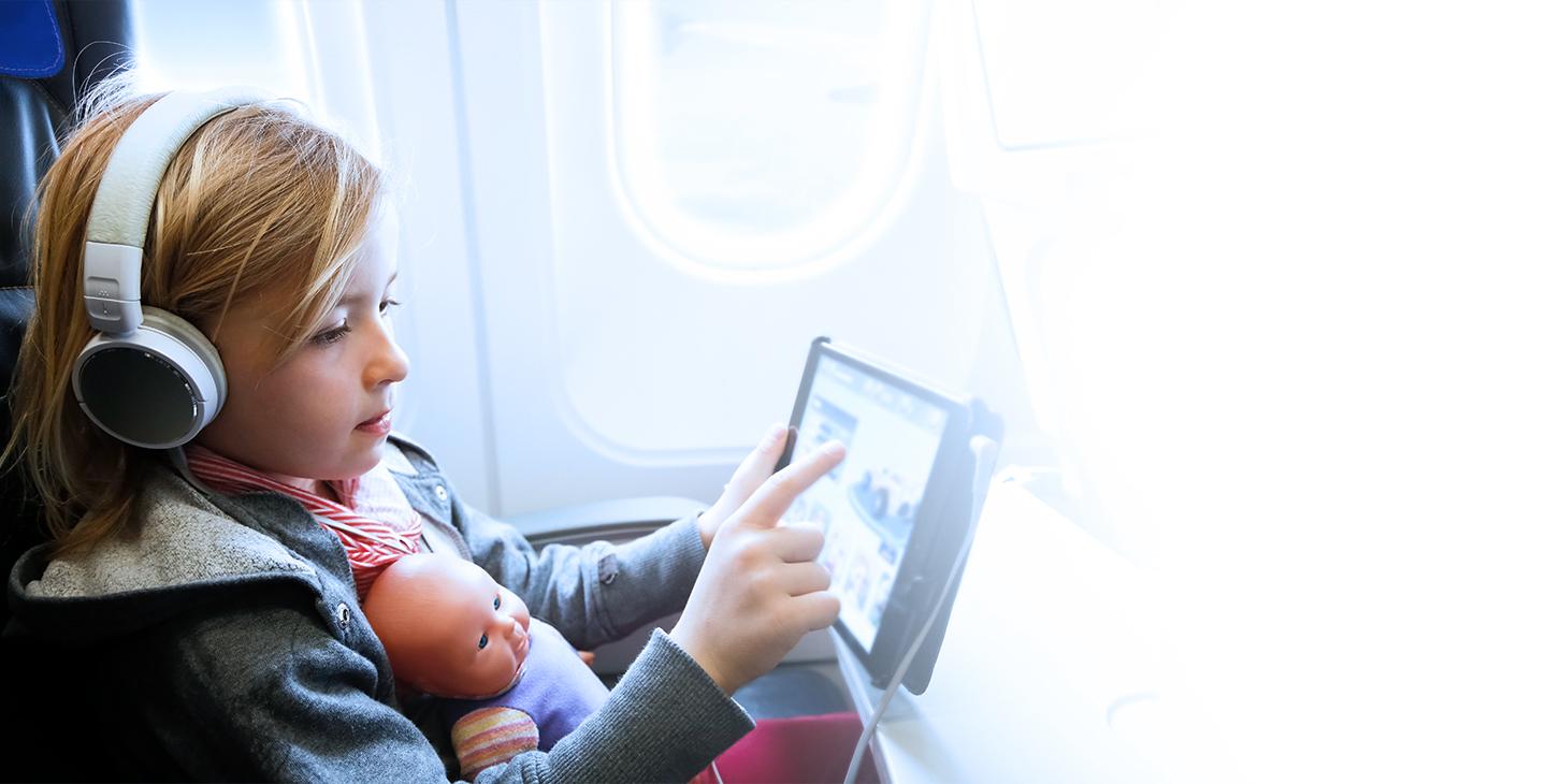 Young girl wearing headphones with a baby doll in her lap enjoying inflight entertainment on her tablet