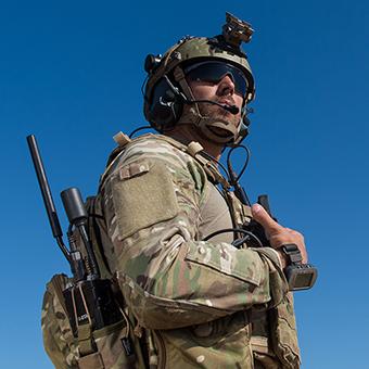 一名身穿绿色迷彩制服的士兵，头戴头盔，戴着太阳镜，背着一个装有收音机的背包